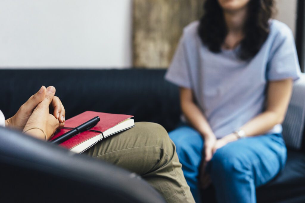 A person has their hands clasped in their lap over a red journal while a young girl sits blurred out in the background attending a therapy session.