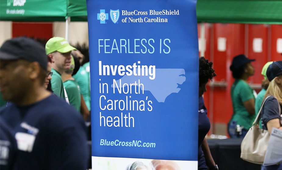 A poster of Blue Cross Blue Shield of North Carolina is centered in front of green tent. People wearing green clothes and hats stand in the background. It reads "Fearless is investing in North Carolina's health".