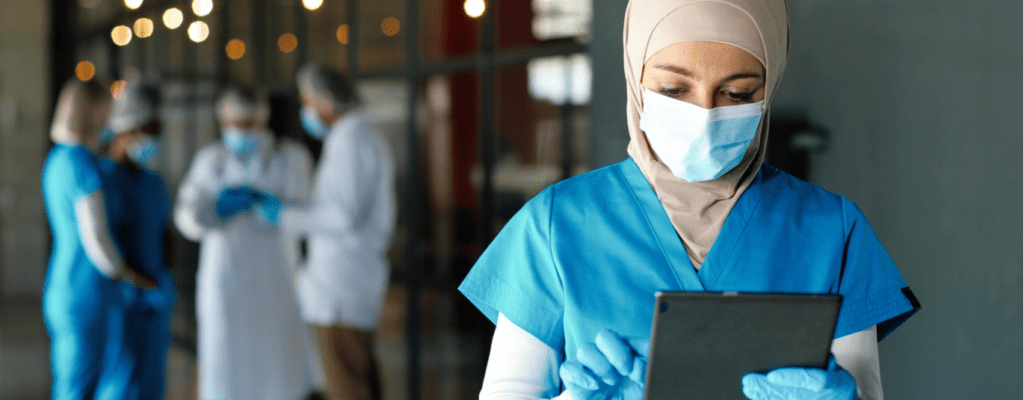 A young woman wearing a hijab, a mask, blue scrubs, and blue rubber gloves looks down at her tablet.