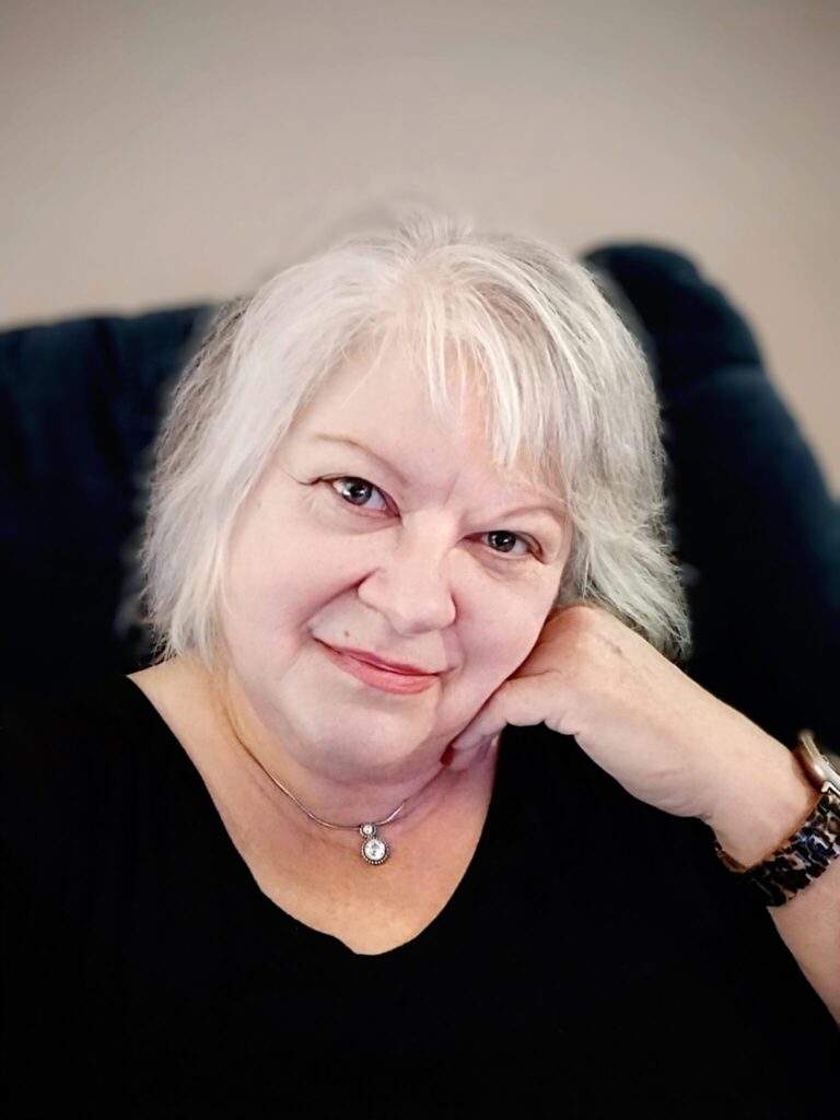 Cindy, an older woman with short white hair, lipstick, and a diamond pendant necklace, smiles at the camera. Cindy is a Customer Service Advocate at BCBSNC.