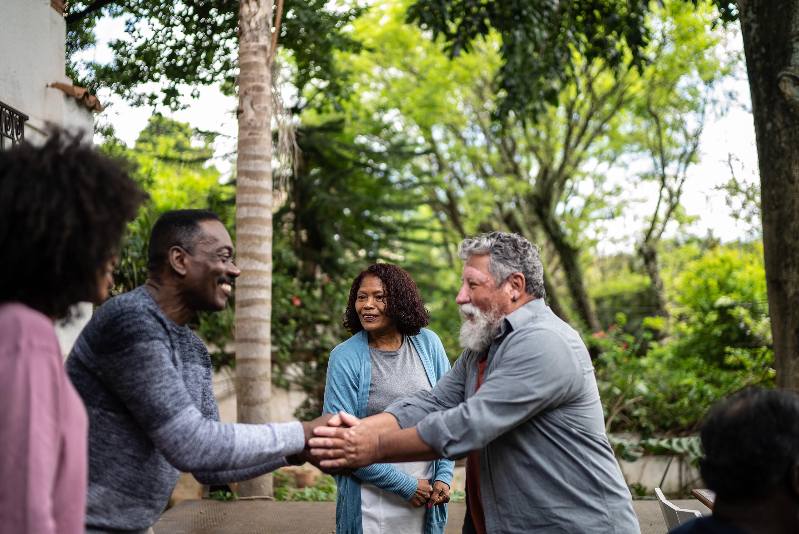 A middle aged African American man stands outside and shakes hands joyously with an older Caucasian man with vitiligo and a long white beard. A middle aged African American woman stands in the background smiling and wearing a blue cardigan.