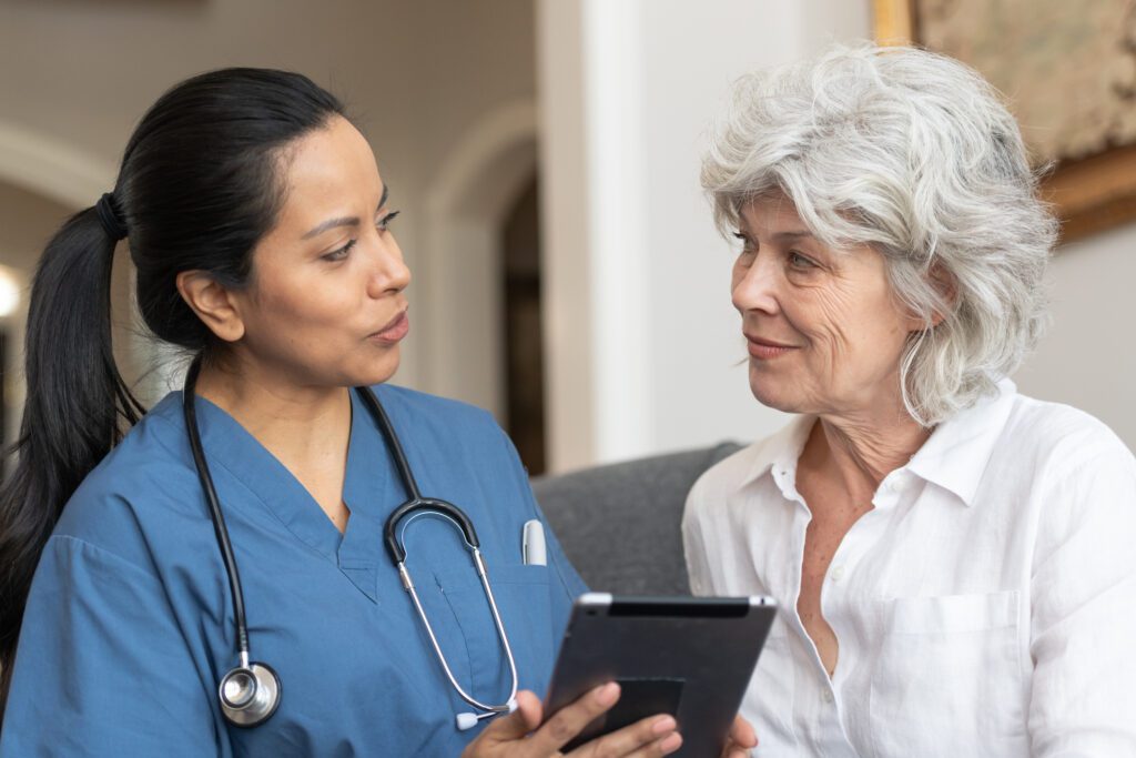 A health care provider wearing blue scrubs and a stethoscope around her neck looks at a tablet with her patient, an older woman wearing a white shirt with vibrant gray hair.