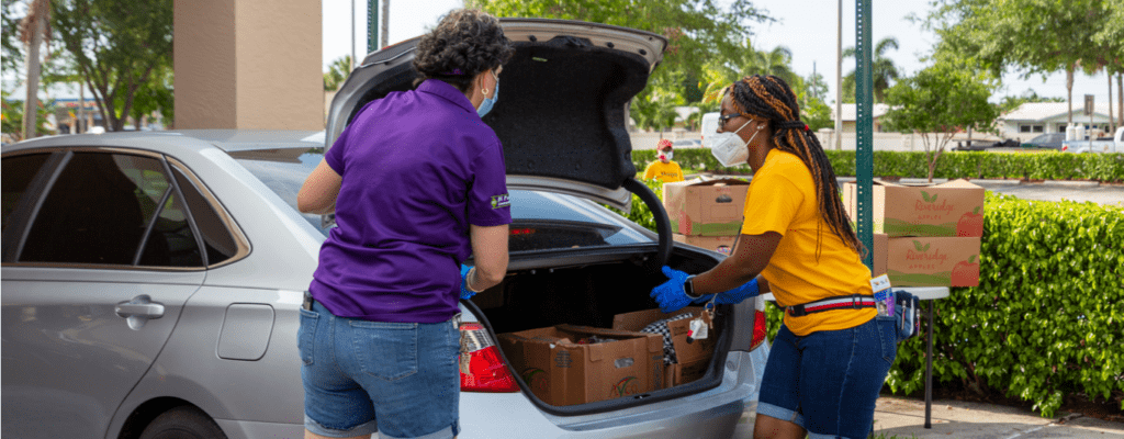 Two women, one with short brown hair in a purple shirt and jean shorts and the other a young African American woman in a bright yellow shirt, blue gloves on her hands, and a mask, stand behind the trunk of a parked car and lift cardboard boxes out of it.