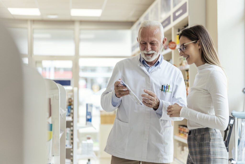 An older man with a white beard smiles while wearing a white lab coat standing in a pharmacy and shares information with a young woman in glasses, long brown hair, a white shirt, and plaid pants.