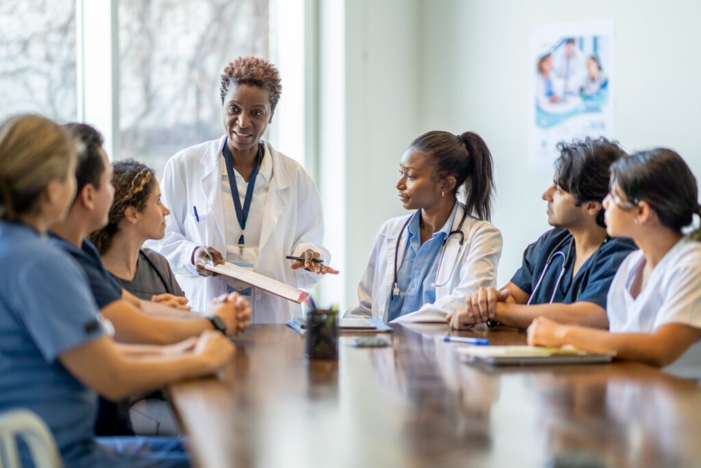 A middle-aged African American female medical instructor stands at the head of a wooden table holding a clip board in front of six students of varying ethnicities.