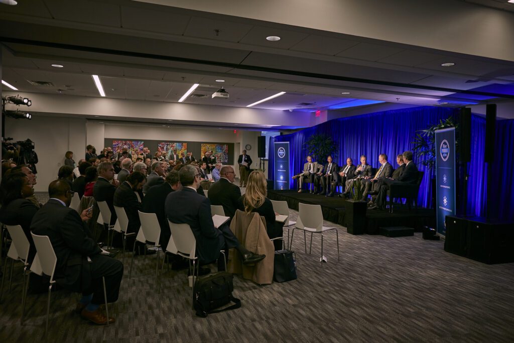 Photo from the Blue Premiere shows people in suits and fancy attire sitting in rows in a conference room looking at a stage where 7 men sit on a panel.