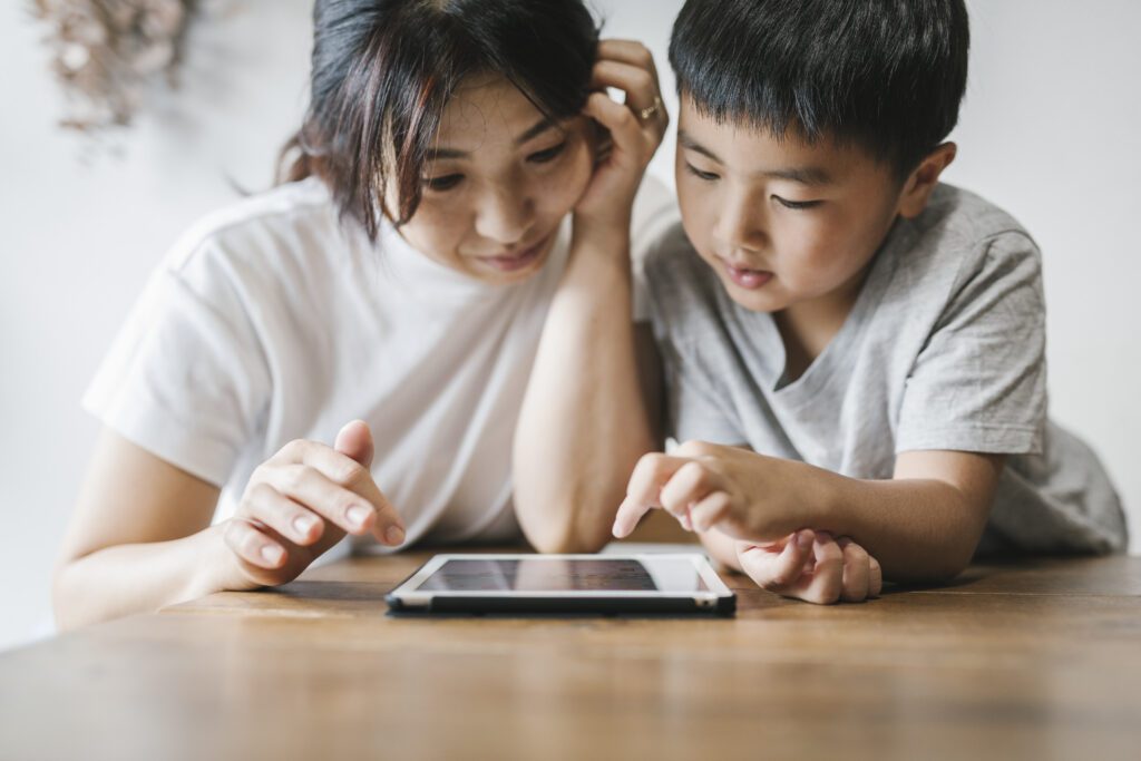 A young Asian mother with black hair wearing a white t-shirt looks at a tablet on a wooden table with her young son.