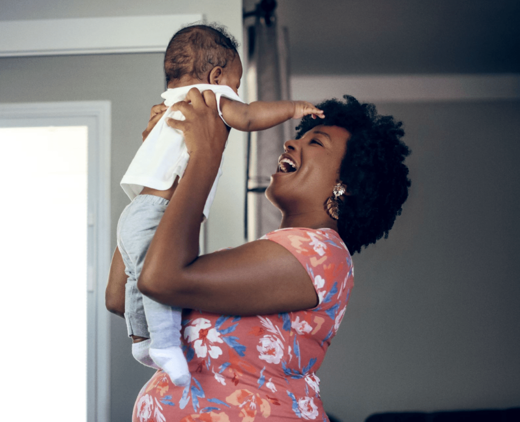 An African American woman with short curly hair and a floral shirt smiles as she holds a young child only a few months old up in the air.