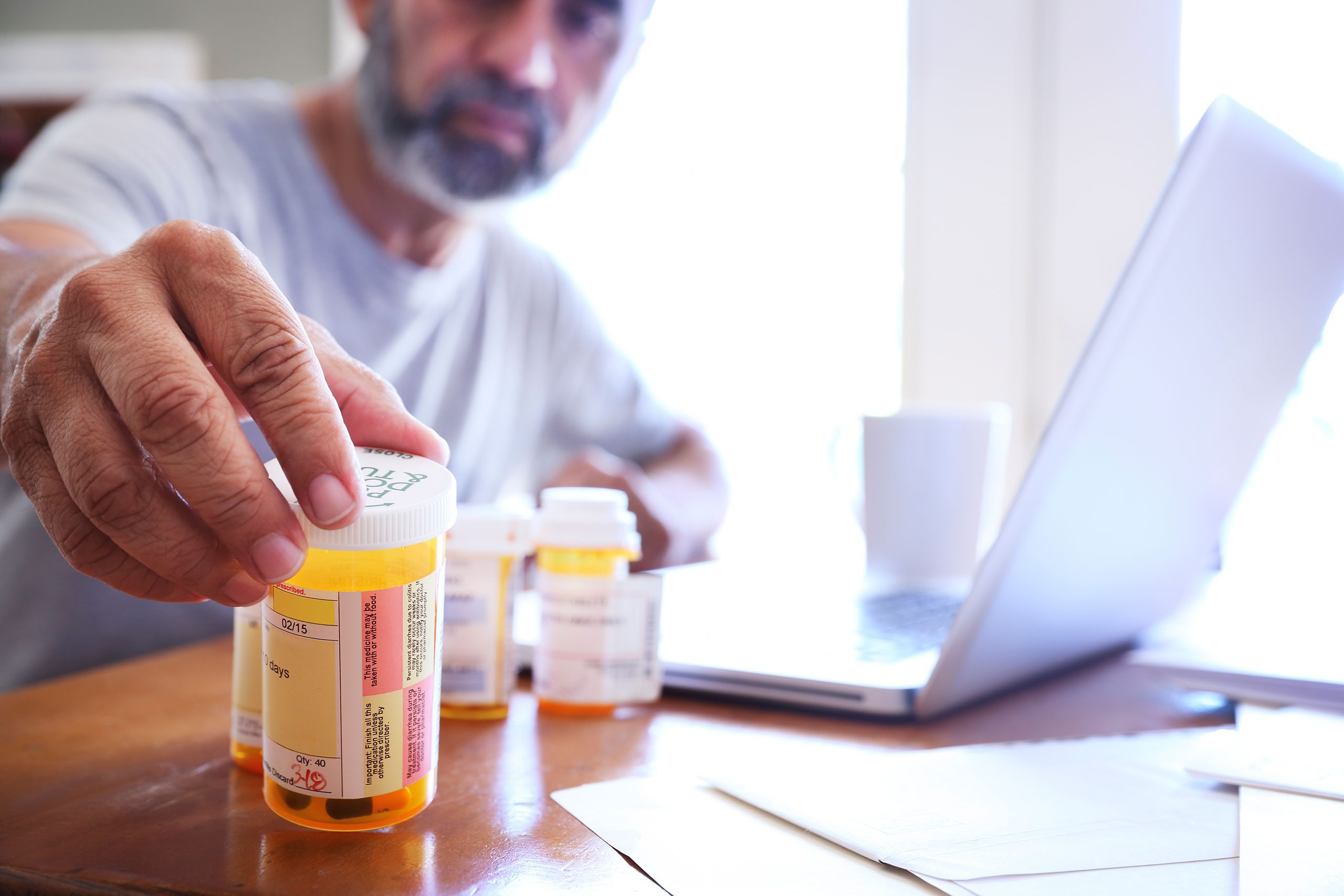 A Hispanic man in his late fifties reaches for one of his prescription medication bottles as he sits at his dining room table. His laptop computer is open in front of him while sunlight filters in through the window behind him bathing the room with a soft glow of light.