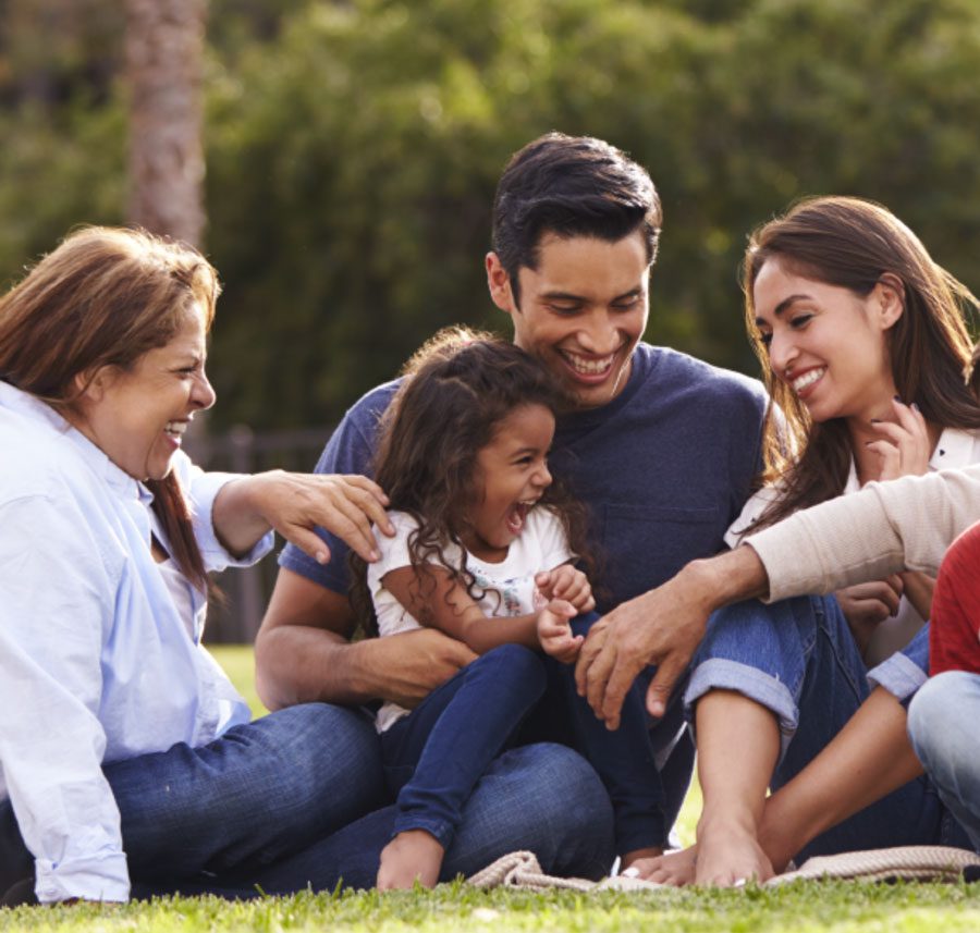 A multiracial family sits together laughing in a park surrounding a young girl in the middle who yells with joy.