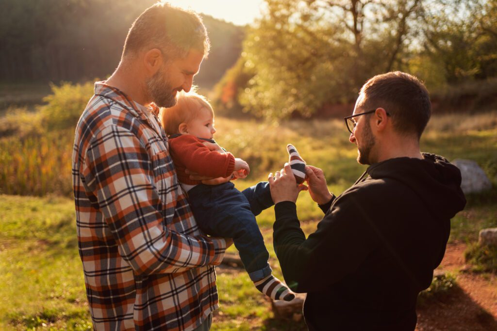 Two men stand outside during sunset. One man in a plaid shirt holds a young toddler while the other man in a dark hoodie and glasses adjusts the toddler's left sock.