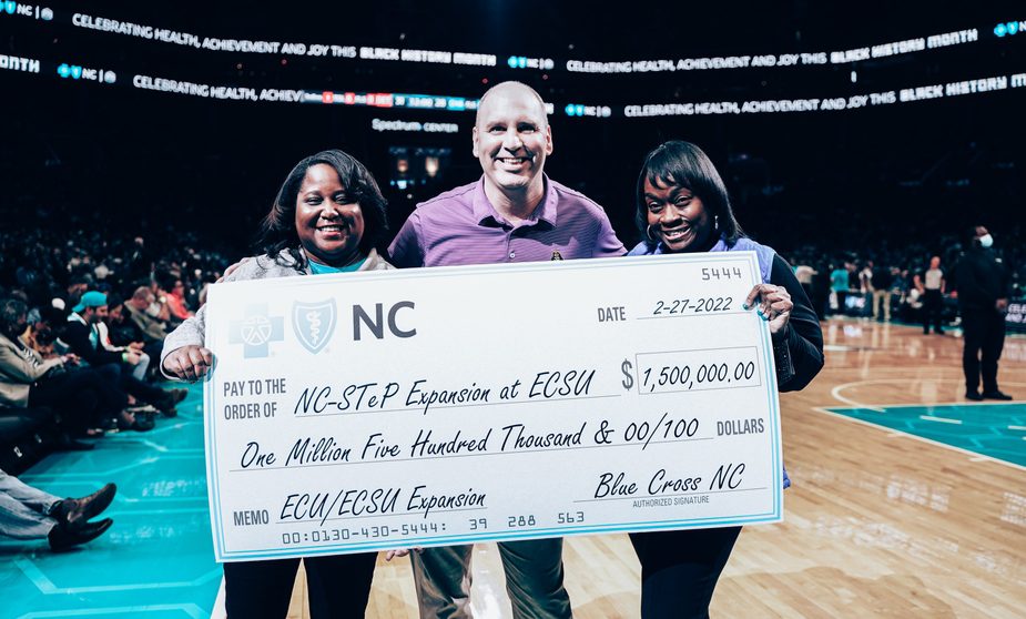 Two young African American women and an older Caucasian man stand smiling holding a large check from Blue Cross Blue Shield made out to Elizabeth City University in North Carolina.