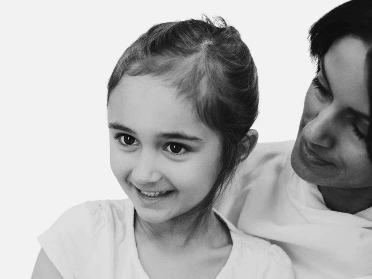 A young girl in a plain T-shirt smiles into the distance and her mother looks at her with care from behind her.