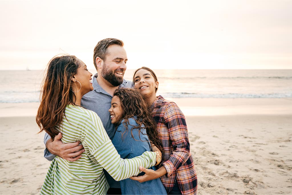 A family of four wearing long sleeve shirts hugging on the beach.