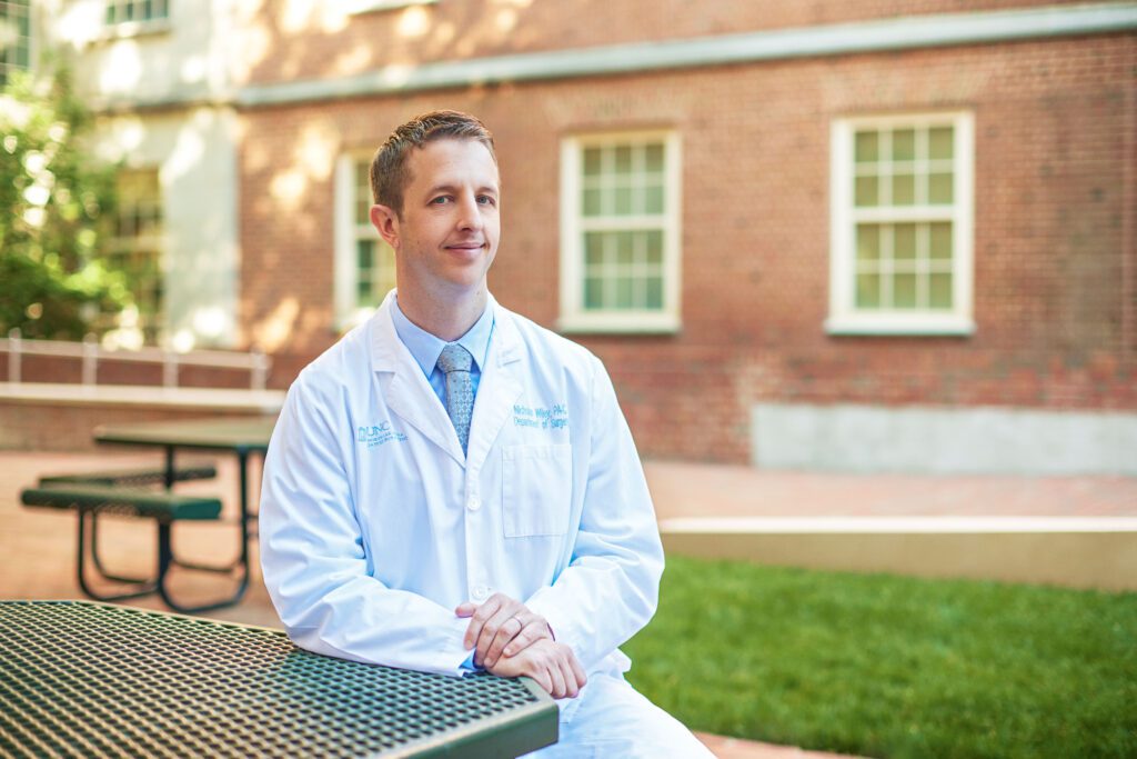 A medical professional in a white lab coat sits at an outside table, looking at the camera
