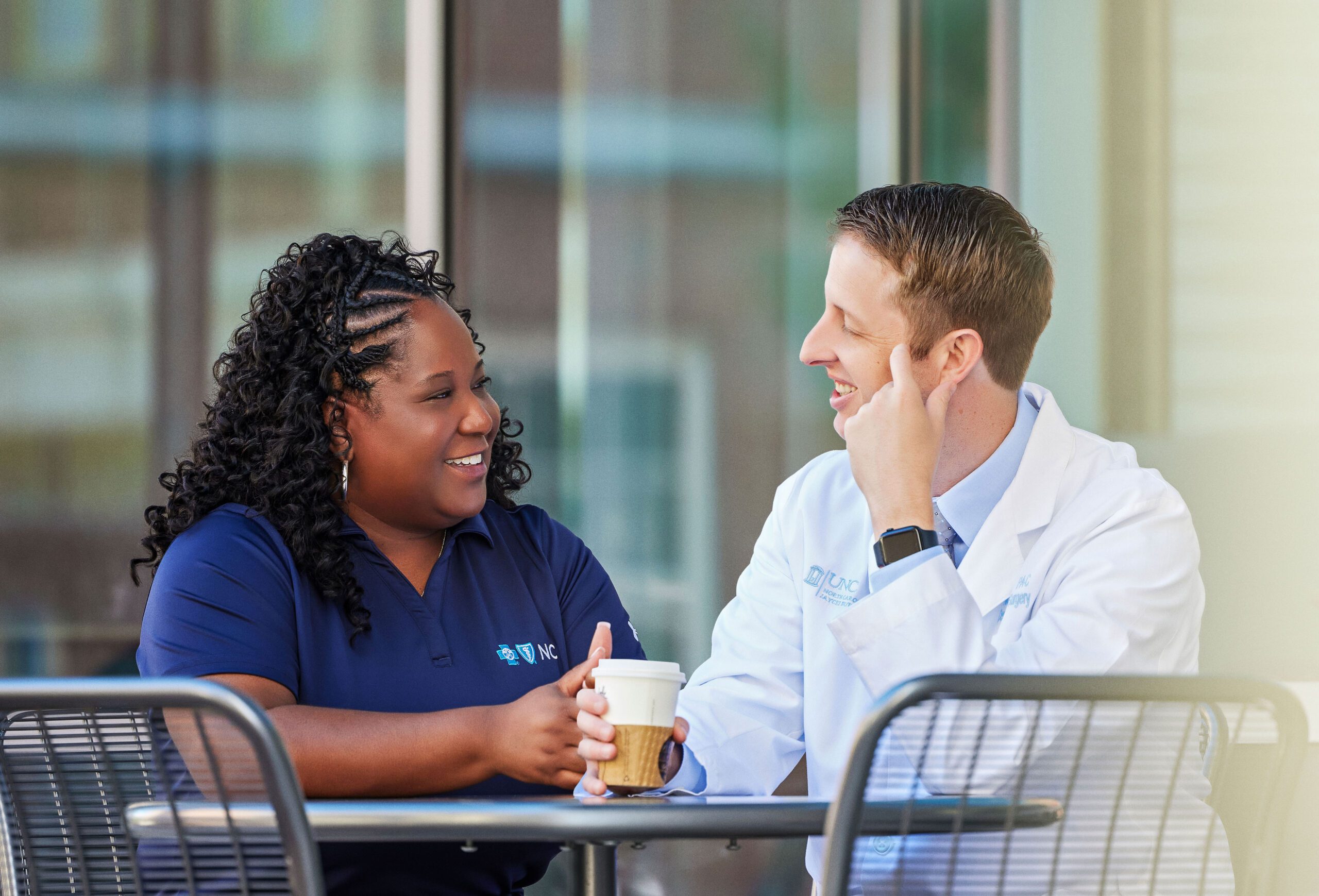 A man in a white lab coat sits an outdoor table, coffee in hand, smiling at a woman seated next to him.